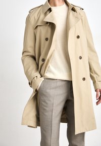 Beige trenčkot s dvojitým zapínáním, velkými hnědými knoflíky a ramenními vycpávkami. Nosený přes krémový svetr a šedé vzorované kalhoty.