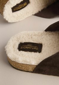 Chaussons en daim marron avec doublure blanche duveteuse et semelles en liège, étiquetés "Next Signature" à l'intérieur.