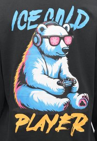 Zwart T-shirt met een kleurrijke afbeelding van een cartoon ijsbeer die roze zonnebril en koptelefoon draagt en een gamecontroller vasthoudt. De tekst luidt "ICE COLD PLAYER."