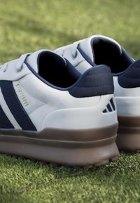 Λευκά και ναυτικό μπλε παπούτσια γκολφ με καφέ καρφιά στη σόλα που ακουμπούν σε πράσινο γρασίδι.