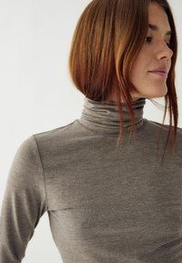 Pelēks, piegulošs turtlenecks tops, izgatavots no mīksta, gluda auduma ar augstu apkakli, garām piedurknēm un minimālu šuvju apdari.
