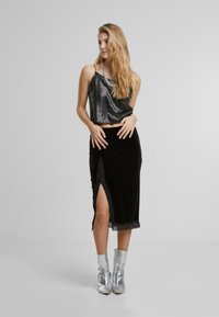 Juoda aksominė midi sijonas su nėriniu apdaila ir šoniniu įpjovimu, derinamas su blizgančia juoda su taškeliais kamizolė. Modelis vilki sidabrinius kulkšninius batus.