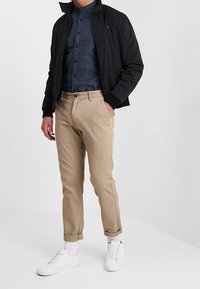 Chaqueta negra ligera, camisa azul marino abotonada, pantalones chinos beige y zapatillas blancas. Texturas suaves y diseño ajustado visibles.