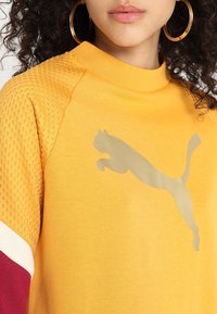 Żółta bluza z teksturowanymi siateczkowymi rękawami, złotym logo Puma i prążkowanym kołnierzem. Ma bordowe i białe akcenty na jednym rękawie.