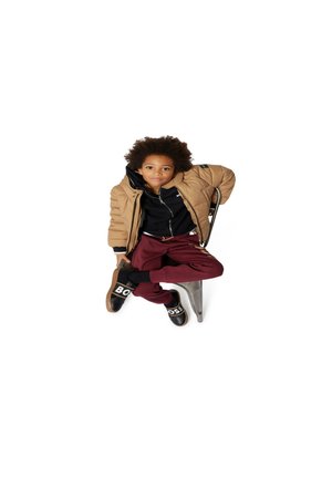Enfant portant une veste en duvet beige, un hoodie noir zippé, un pantalon bourgogne et des baskets noires avec le logo blanc "BOSS", assis sur une chaise en métal.