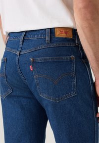 Los pantalones vaqueros de denim azul oscuro presentan un diseño de bolsillo trasero distintivo con una etiqueta roja, remaches de cobre y un patrón de costura amarilla.