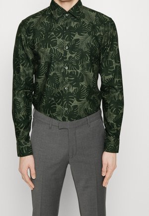 Homme portant une chemise à boutons vert foncé avec un motif de feuilles et un pantalon de costume gris sur fond blanc.
