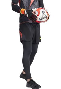 Pantaloni sportivi neri con accenti arancioni e verdi, abbinati a leggings neri e scarpe da calcio. La figura tiene in mano un pallone da calcio rosso e bianco.