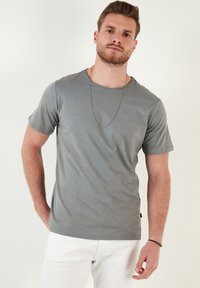 Šedé tričko s krátkým rukávem z hladkého materiálu, s klasickým výstřihem, jemnou texturou a volným střihem. Nosené s bílými kalhotami.