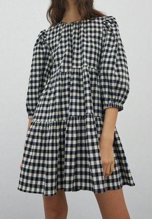 Woman wearing a black and white checkered dress with puffed sleeves and a tiered skirt, standing against a plain light background.