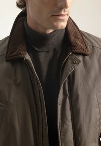 Chaqueta acolchada marrón con un cuello de pana marrón, combinada con un suéter de cuello alto oscuro. Cierre frontal con cremallera y detalles de hardware visibles.