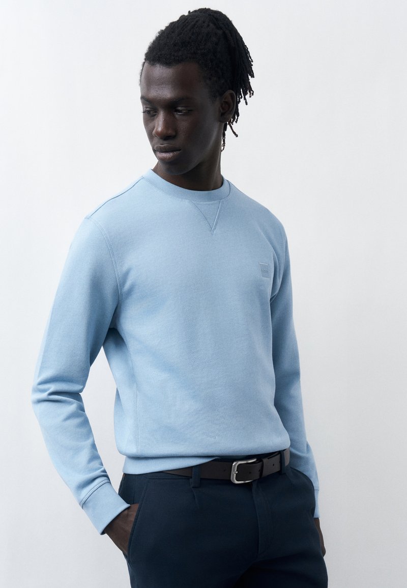 Νέος άνδρας με σκουρόχρωμο δέρμα και ράστα, φοράει ανοιχτό μπλε φούτερ και σκούρο παντελόνι, με τα χέρια στις τσέπες, κοιτάζει προς τα κάτω πλάγια.