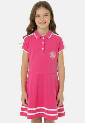 Fille aux longs cheveux bruns portant une robe polo rose avec des bordures blanches et un petit logo rond sur la poitrine, souriant à la caméra.