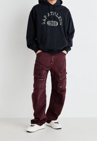 Sudadera negra con el gráfico "GAP ATHLETIC 1969", combinada con pantalones cargo granate que tienen múltiples bolsillos y un detalle de cremallera, usados con zapatillas blancas.