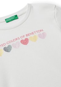 Bílý bavlněný tričko s žebrovanou texturou, s designem srdce v růžové a třpytivé barvě. Nápis "United Colors of Benetton" je vytištěn růžovou barvou nad srdíčky.