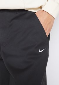 Černé látkové kalhoty s boční kapsou, jemnou texturou a bílým logem Nike vpředu blízko kapsy.