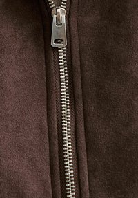 Tela marrón con una textura suave, que presenta un zipper plateado en el centro. El zipper tiene un tirador rectangular y una pista suave.