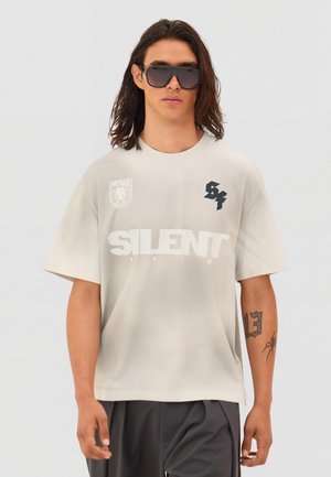 Mladić s dugom kosom, koji nosi velike sunčane naočale, bež majicu s natpisom „SILENT“ i crne hlače na jednobojnoj sivoj pozadini.