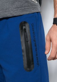 Pantaloni sportivi blu con una tasca laterale con zip nera e brandizzazione. Il tessuto appare morbido e con una texture liscia.