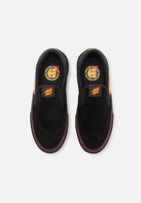 Melnas zamšādas slip-on kurpes ar sarkaniem akcentiem, ar noapaļotu purngala formu un logo uzlīmēm uz mēles un zoles. Tekstūrēta augšdaļa ar šuvju detaļām.