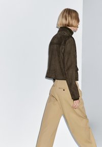 Bruine cropped suède jas met een hoge kraag, gedragen over een donker gebreide trui, gecombineerd met losse beige broek met achterzakken en zwarte knopen.