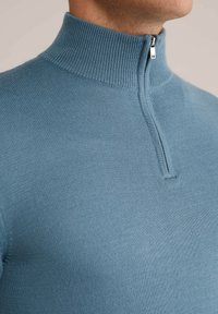 Heled sinine tõmbluku kaelusega sviiter, mis on valmistatud tekstureeritud kudumifaasist. Omab kõrget kaelust ja lühikest tõmblukku. Üksikasjalik ja lähedalt istuv siluett.