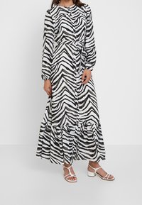 Lång skjortklänning med svartvit zebra-mönster, långa ärmar och en flerlagrad volang längst nere. Bärs med vita sandaler med remmar.