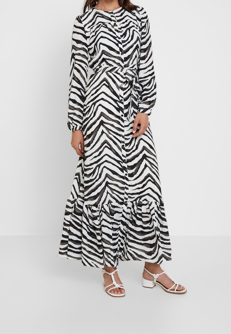 Lång skjortklänning med svartvit zebra-mönster, långa ärmar och en flerlagrad volang längst nere. Bärs med vita sandaler med remmar.