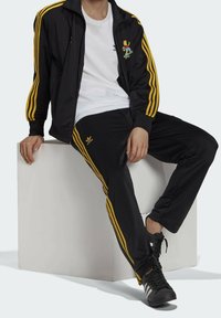 Svart idrottsbyxdress med gula ränder, med en tecknad design på jackan. Bärs över en vit t-shirt, ihop med svarta sneakers.