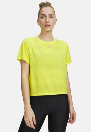 Vrouw die een felgeel, korte mouwen, mesh sportshirt draagt en zwarte legging, staand tegen een effen lichte achtergrond.