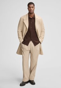 Cappotto di lana beige, camicia marrone a bottoni, pantaloni beige a righe sottili e stivali neri. L'outfit presenta una vestibilità rilassata e dettagli sartoriali.