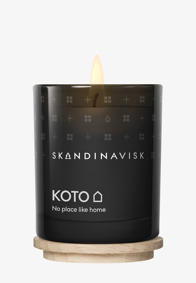 Svart glasljushållare med dekorativa mönster, träbas och en tänd veke inuti. Texten inkluderar "SKANDINAVISK KOTO Ingen plats som hemma."