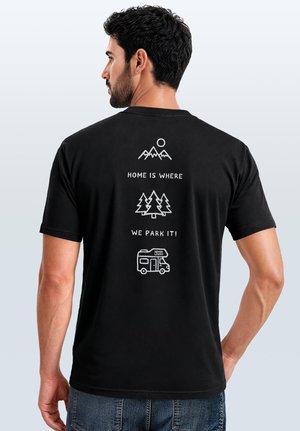 Mann trägt schwarzes T-Shirt mit weißem Design: Berge, Bäume und Wohnmobil mit dem Text „Home is where we park it!“ auf dem Rücken.