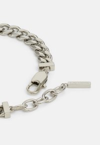 Hõbedane kett käevõru suurte ovaalsete linkidega, koos homaariklambriga ja ristküliku kujulise sildiga, millel on embosseeritud tekst "LACOSTE". Sile metalliline tekstuur.