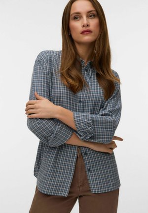 Femme aux longs cheveux bruns portant une chemise à carreaux bleue et grise avec des boutons et un pantalon marron, se tenant bras croisés sur un fond blanc.