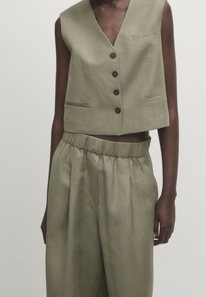 Veste sans manches vert olive à boutons, associée à un pantalon large taille haute avec ceinture élastique assorti, portée par une personne à la peau foncée.