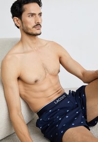 Navy blue boxer shorts with a white Lacoste logo, featuring a repeated light blue crocodile pattern, made of lightweight fabric.