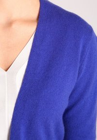 Blå stickad kofta med V-ringning, gjord av mjukt material. Har en sömlös textur och bärs över en ljusfärgad topp.