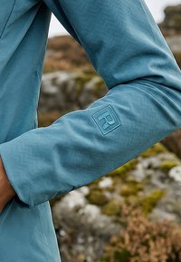 Jachetă bleu cu mâneci lungi și siglă pătrată în relief pe brațul superior, purtată în aer liber, pe fundal natural estompat cu stânci.