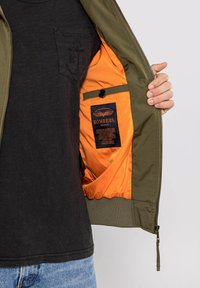 Oliwkowa bomberka z pomarańczową podszewką, zapinana na zamek, z czarną metką. Posiada kieszeń wewnętrzną oraz ściągacze na rękawach. Nosi się ją na ciemnym t-shircie.