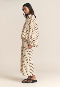 Beige top met stippen, losvallend met hoge kraag, gecombineerd met bijpassende rok met stippen. Zwarte slippers maken de outfit compleet.