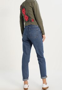 Khaki skórzana kurtka typu cropped z czerwonym haftem kwiatowym z tyłu, w połączeniu z niebieskimi dżinsami z podwyższonym ściągaczem i białymi trampkami. Casualowy, luźny krój.