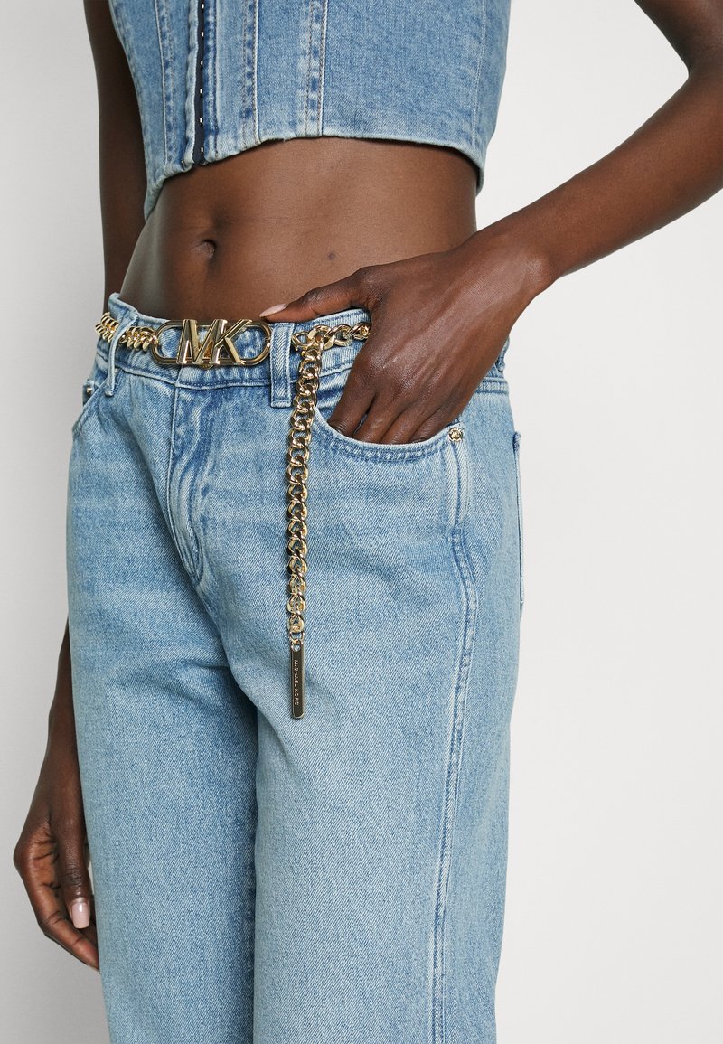 Jasnoniebieskie dżinsy z złotym okuciem i paskiem łańcuszkowym z emblematem "MK", udekorowane złotą pętlą łańcucha.