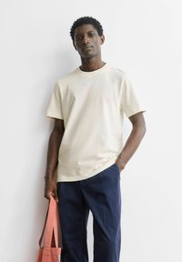 Beige katoenen T-shirt met korte mouwen en een ronde hals, gecombineerd met marineblauwe broek. Het model houdt een roze stoffen tas vast.
