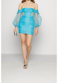 Tyrkysové šaty bez ramínek s průhlednými nabíranými rukávy, rýhovanou texturou a bočními výřezy. Spárované s chromovou kabelkou a metalickými sandály.