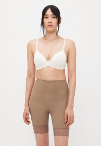 Vrouw met zwart haar die een witte beha en hoge taupe fietsshorts met kant draagt, staande tegen een effen witte achtergrond.
