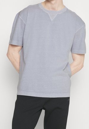 Man draagt een lichtgrijs shirt met korte mouwen en zwarte broek, staand met handen achter de rug, tegen een effen lichte achtergrond.