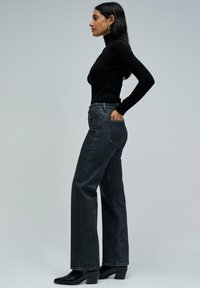 Un cuello de tortuga negro combinado con jeans de mezclilla oscuros de cintura alta. Los jeans tienen un corte holgado y bolsillos de parche; los zapatos son tacones negros.