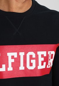 Felpa con corpo nero e una vistosa striscia orizzontale rossa. "ELFIGER" stampato in bianco, con "DENIM" in caratteri più piccoli. Texture morbida.