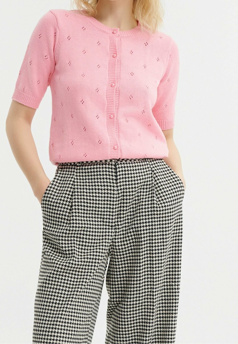Femme portant un cardigan rose à manches courtes boutonné avec un motif de petits trous et un pantalon pied-de-poule noir et blanc taille haute, les mains dans les poches.